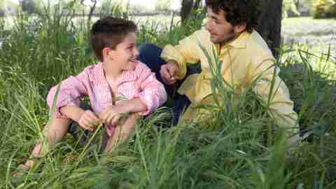 A father sits in the grass under a tree and talks with his son about executive function