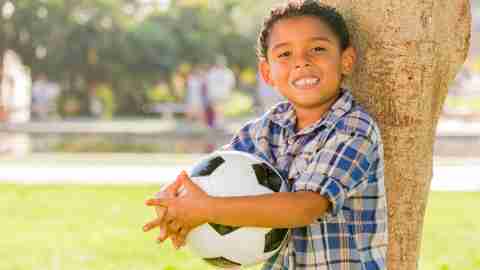 A boy with ADHD holds a soccer ball and leans against a tree in a park