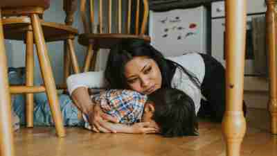 A little boy lies face down on the floor under a table, while a woman comforts him