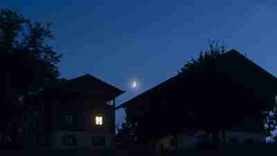 Illuminated window on rural house with crescent moon at blue hour. Image used to convey delay sleep phase syndrome, or DSPS, a condition that causes individuals to have difficulty falling asleep at socially conventional times. DSPS disproportionately affects individuals with ADHD and depression.