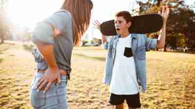 Preteen boy is smirking and holds a skateboard against his shoulders. His frustrated mother stands before him.